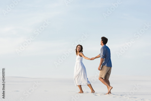 young couple walking on sandy beach and holding hands against blue sky