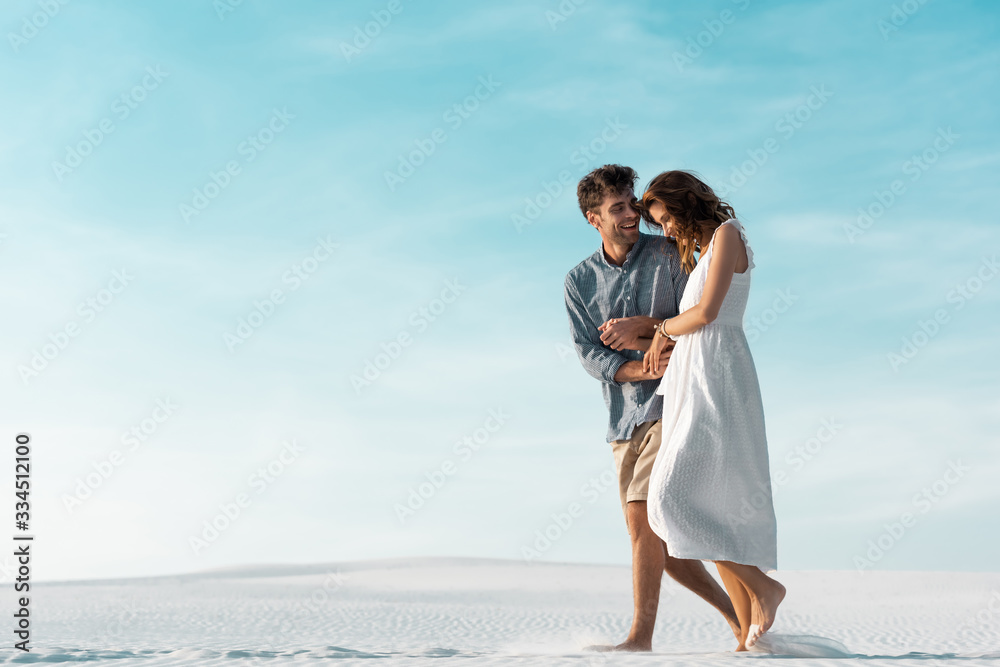 smiling young couple walking on sandy beach against blue sky