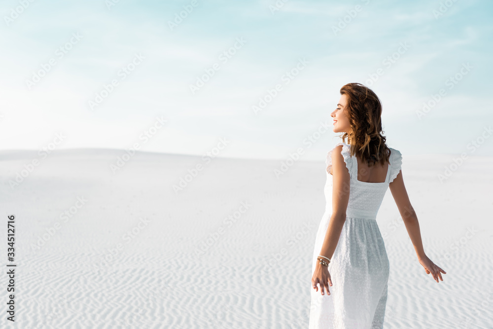 back view of smiling beautiful girl in white dress on sandy beach with blue sky back view of smiling beautiful girl in white dress on sandy beach with blue sky