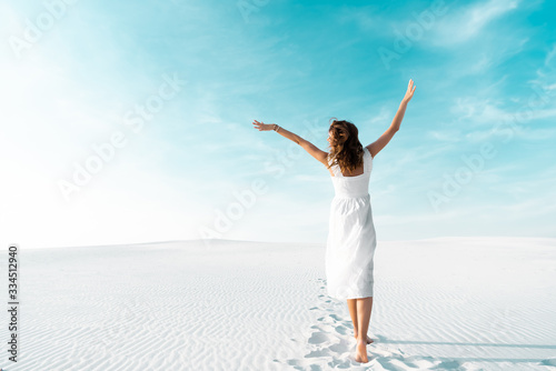 back view of beautiful girl in white dress with hands in air on sandy beach with blue sky