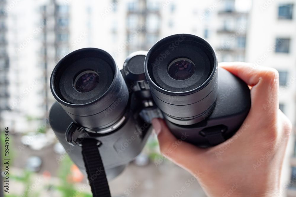 Hand holding black binoculars against a blurred background of a window with a view of the neighboring house for observing neighbors, environment or nature. Selective focus. Closeup view