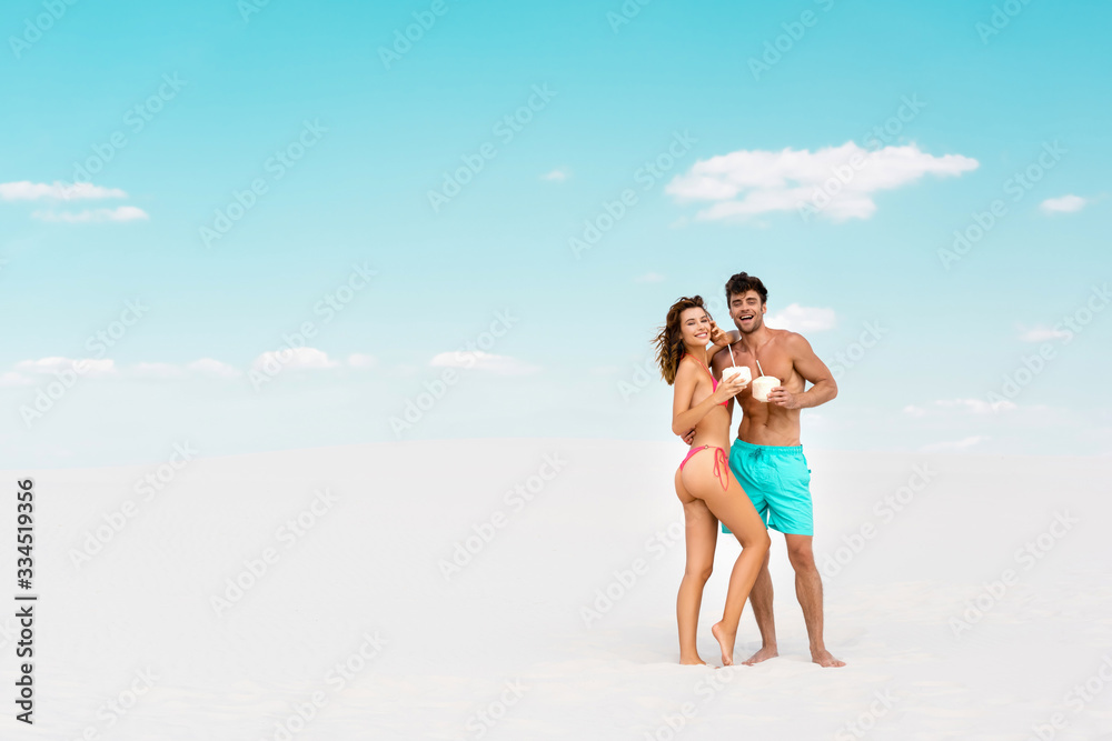 smiling young couple embracing while holding coconut drinks on sandy beach