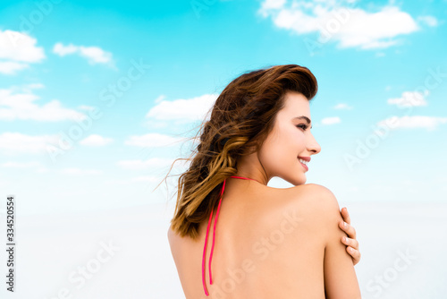 back view of smiling beautiful sexy girl in swimsuit on beach with blue sky and clouds