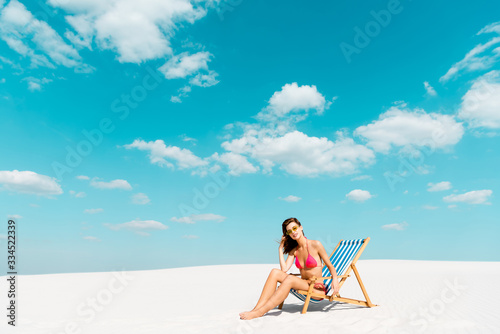beautiful sexy girl in swimsuit and sunglasses sitting in deck chair on sandy beach with blue sky and clouds © LIGHTFIELD STUDIOS