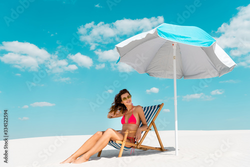 smiling beautiful sexy girl in swimsuit and sunglasses sitting in deck chair under umbrella on sandy beach