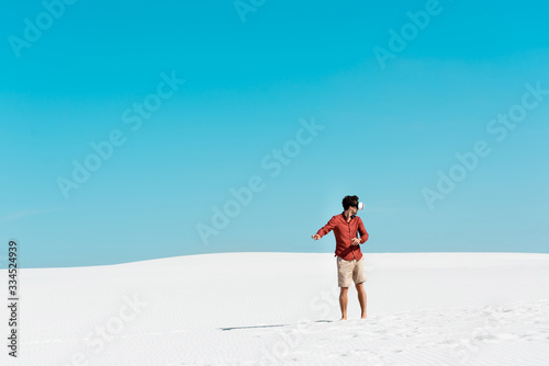 man on sandy beach in vr headset gesturing against clear blue sky