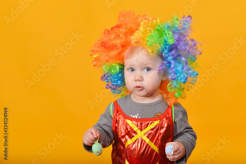 A small child, a girl in a clown wig, holds Easter eggs in her hands.