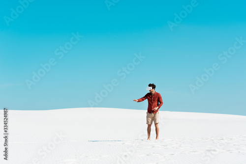 man on sandy beach in vr headset gesturing against clear blue sky