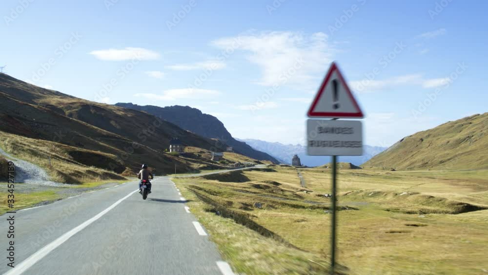 Male man on a motorbike crossing french border sign on a scenic mountain pass road surrounded by the green fields with blue sky and some clouds. Camera from behind.