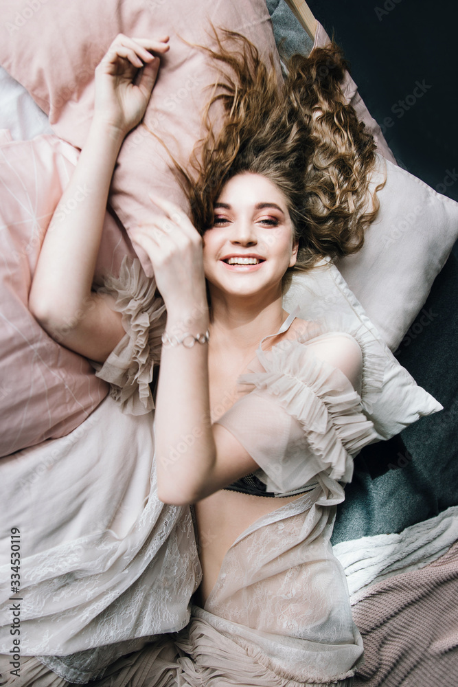 The smiling girl in a transparent dressing gown lies on a bed with pink linen, top view