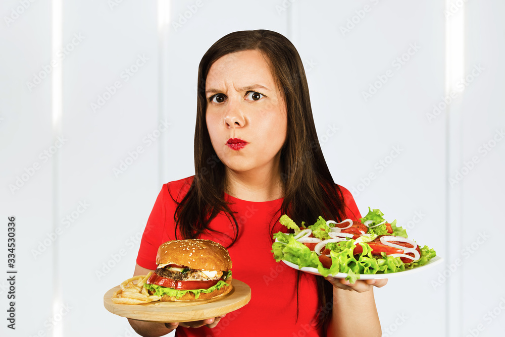 Young woman with excess weight holds hamburger and plate of salad Young woman with excess weight holds hamburger and plate of salad