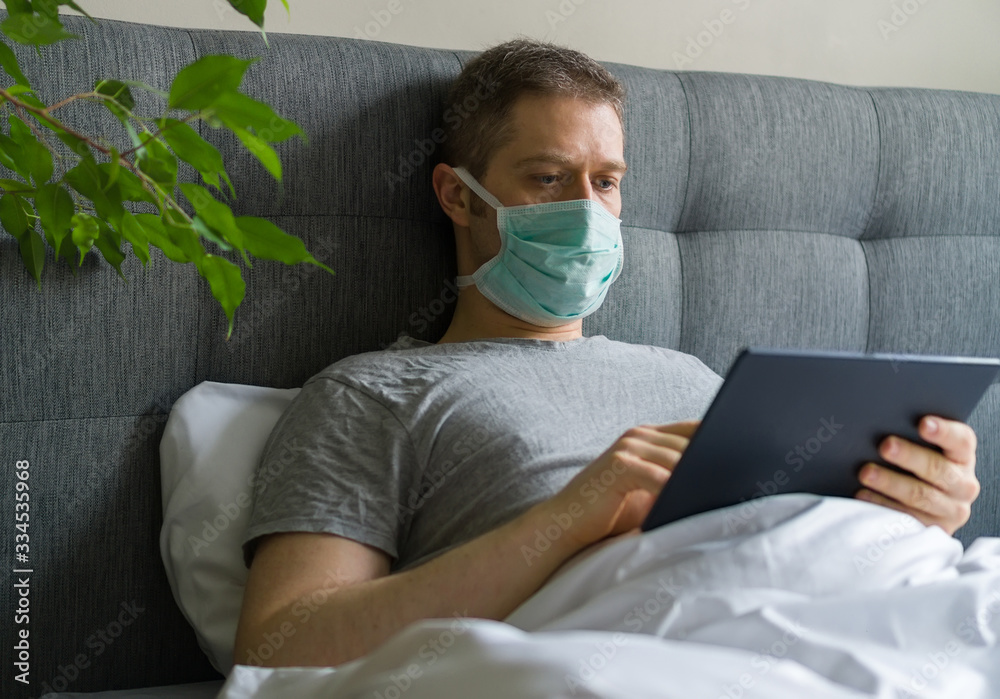 Sick man with medical mask using tablet pc in bed. Home quarantine concept. Sick man with medical mask using tablet pc in bed. Home quarantine concept.