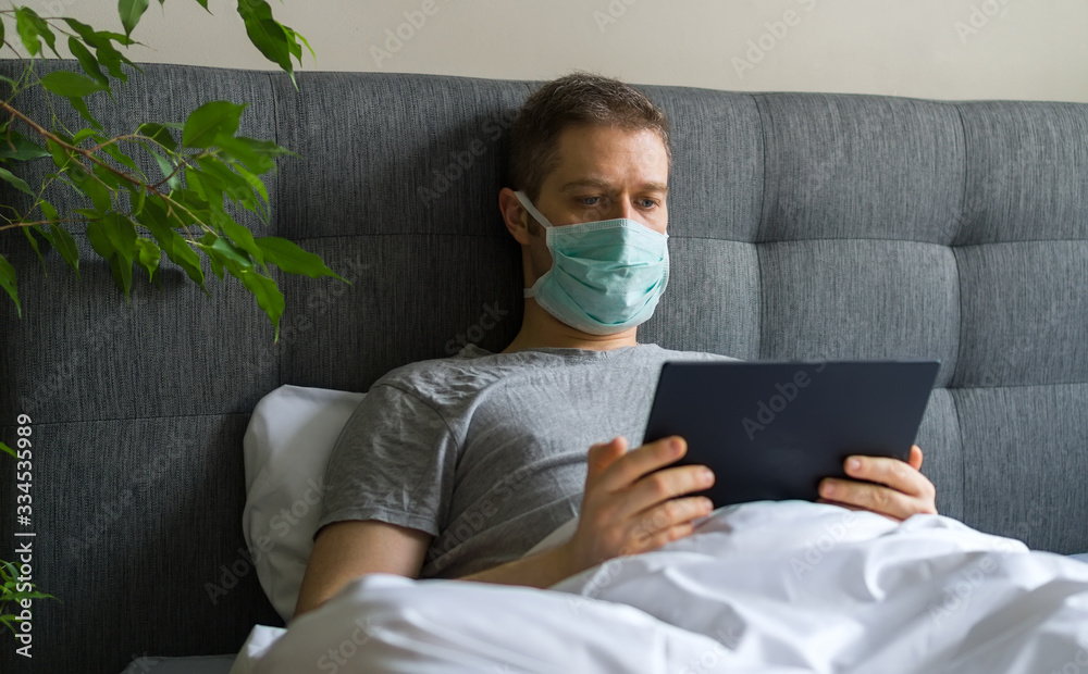 Sick man with medical mask using tablet pc in bed. Home quarantine concept.
