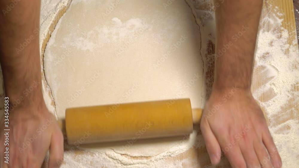 Top view, male hands roll out the dough with a rolling pin on a wooden surface, close-up. Close up of baker or chef preparing fresh dough with rolling pin on kitchen table. Male hands rolling dough.