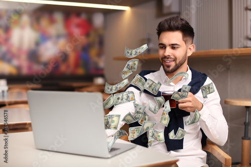 Man with modern laptop and flying dollar banknotes at table indoors. People make money online photo