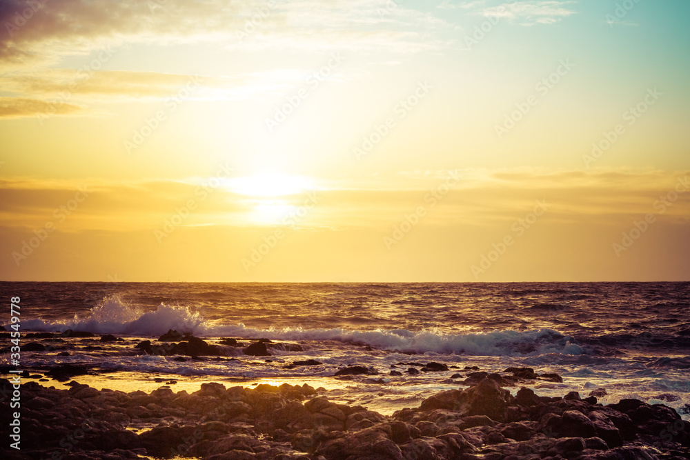 The sun rising over the ocean with a rocky foreground and rough surface crashing into the shore of Hawaii.