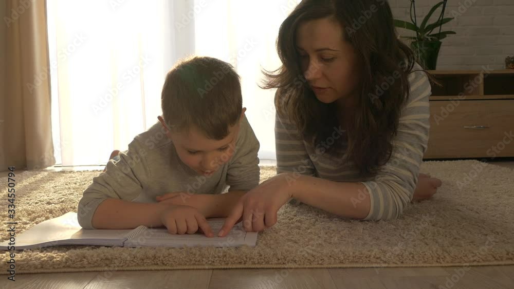 Mom and son are reading a book. Little boy with mom do homework. Reading together is fun at home on the rug.