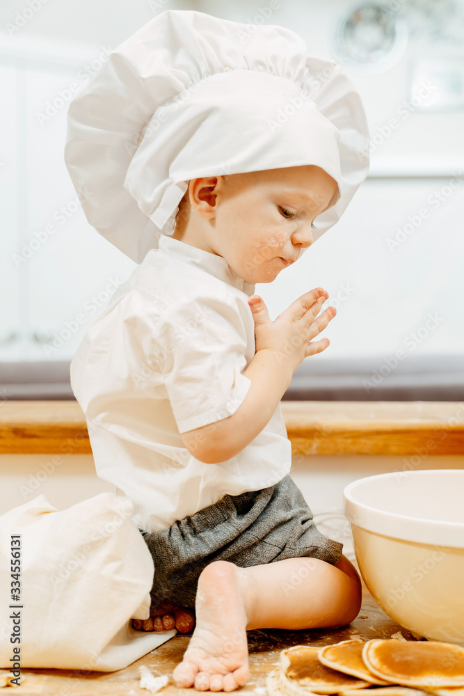 Side view of a naughty little boy in white cook suit sitting on the kitchen table barefoot and kneading dough for fritters. Concept of natural curiosity of children Side view of a naughty little boy in white cook suit sitting on the kitchen table barefoot and kneading dough for fritters. Concept of natural curiosity of children