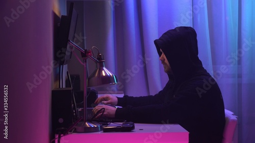 Male hacker sits at his desk and breaks into the system. Man in black hoodie hides his face and works at laptop at neon lighting.