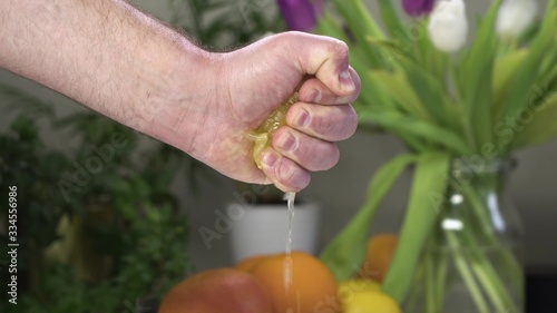 Male hand squeeze lemon with one hand. On background there are fruits and plants.