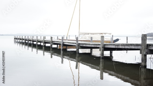 Obraz na płótnie Sailing Boat docking on a pier at a lake with calm water reflection on a grey and cloudy day w salonie