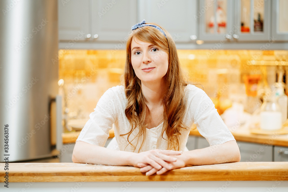 Portrait of a positive young and lovely housewife woman leaning against a wooden bar counter against the backdrop of a high-tech kitchen. Concept of cooking at home Portrait of a positive young and lovely housewife woman leaning against a wooden bar counter against the backdrop of a high-tech kitchen. Concept of cooking at home