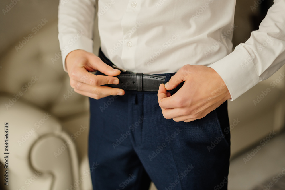 Groom in blue pants and a white shirt fastens his leather belt. Morning groom. Wedding details. Concept and composition. Businessman man and business. Groom in blue pants and a white shirt fastens his leather belt. Morning groom. Wedding details. Concept and composition. Businessman man and business.