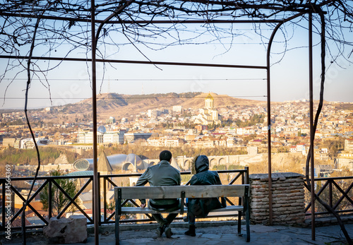 Couple in warm clothes are sitting on the bench and enjoying the view of Holy trinity cathedral and city view. Tbilisi.Georgia.2020