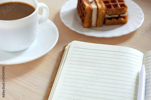 Side view  a cup of fresh coffee  waffles with cream filling on a plate  a notebook are on the table