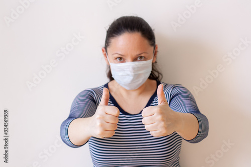 Girl in a medical mask shows thumbs up. European appearance. Long black hair. Focus on outstretched arms  face blurry. Horizontal.