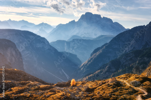 Mountain canyon lighted by bright sunbeams at sunset in autumn. Dolomites, Italy. Landscape with mountain ridges, rocks, colorful trees with orange leaves, alpine meadows, blue sky, sunlight in fall