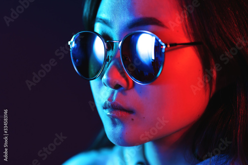 Young pretty girl with asian appearance in neon pink and blue lights at night. Serious trendy hipster teenager in glasses