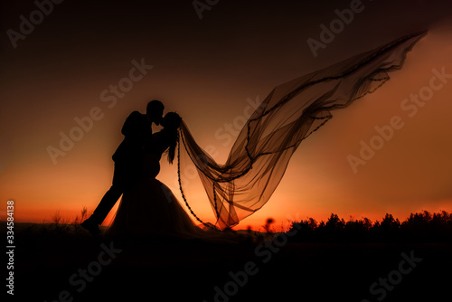 Silhouette of bride and groom kissing at sunset. The veil is flying.