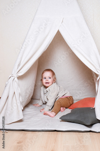 A child, a boy, is sitting and playing in a tent inside the house on a wooden background.