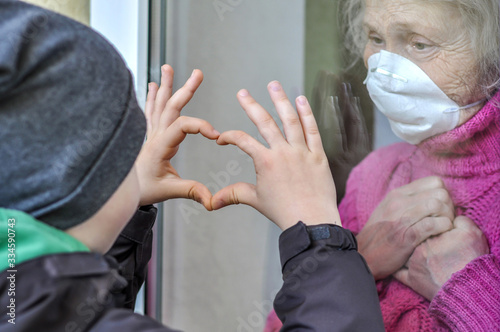 Grandmother mature woman in a respiratory mask communicates with her grandchild through a window. Elderly quarantined, isolated. Coronavirus covid-19. Caring with older people. Family values, love