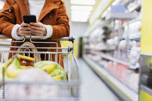 Woman with smart phone pushing shopping cart in supermarket