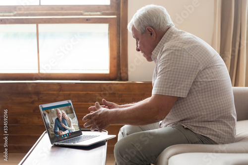 Elderly man sit on couch at home talk on video call on laptop with family friends, senior grandfather relax on sofa in living room speak communicate online use Webcam conversation on modern computer