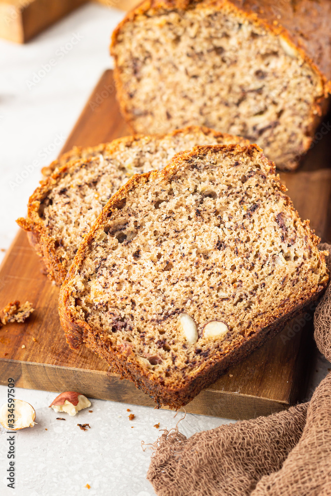 Homemade banana bread or cake with hazelnut on wooden cutting board, light stone background. Trendy soft shadows, atmospheric lighting, selective focus. Flat lay composition. Homemade banana bread or cake with hazelnut on wooden cutting board, light stone background. Trendy soft shadows, atmospheric lighting, selective focus. Flat lay composition.