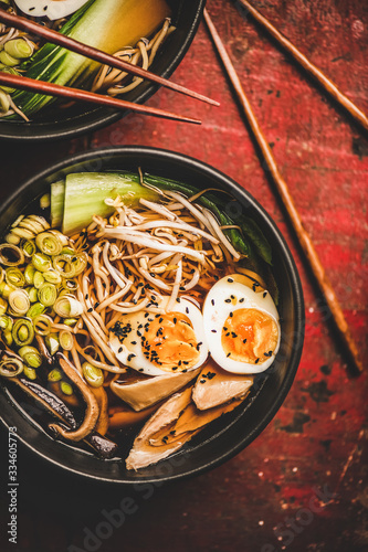 Flat-lay of traditional japanese Ramen soup with chicken meat and shiitake mushrooms in black bowls and bamboo chopsticks over dark red wooden table background, top view. Japanese cuisine concept