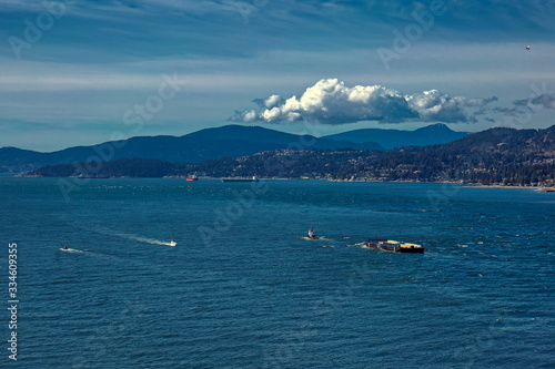 Vancouver harbor, tug-boat tows a barge, ocean tankers are waiting for loading in the port on a sunny windy day against the backdrop of a mountain ridge and cloudy sky