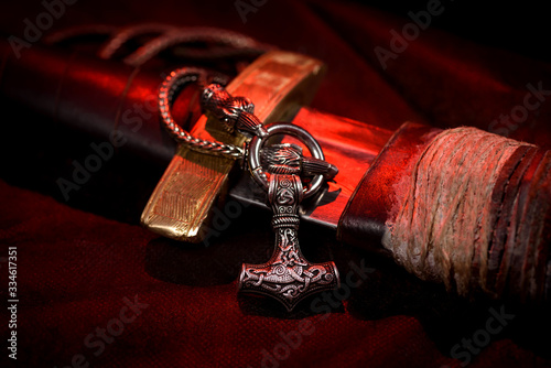 Viking battle sword made of steel with a leather handle with a Thor's silver Hammer pendant on a red black background. Handmade. Mjolnir. Viking's symbol