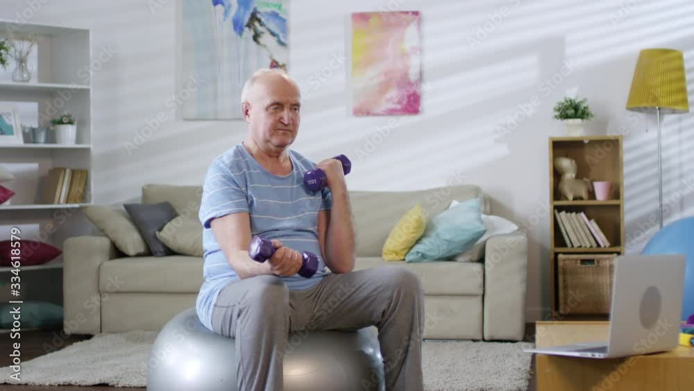 Senior man sitting on exercise ball, watching video workout on laptop and doing dumbbell curls while working out in the living room at home Senior man sitting on exercise ball, watching video workout on laptop and doing dumbbell curls while working out in the living room at home