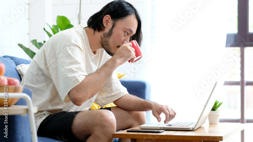 Work from home, social distancing, Asian man working online with laptop computer at home office, Asia male smiling and holding coffee cup at home office , Guy study online education, quarantine people