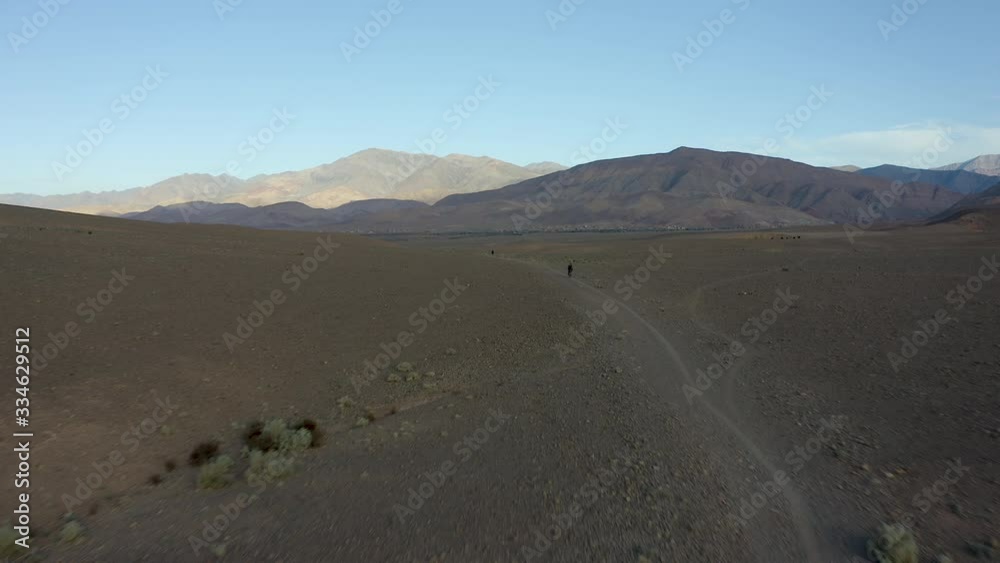 A low aerial flyover of two cyclists bikepacking away from a village at dawn or dusk, across a desolate, rocky desert landscape dotted with sage and rimmed with colorful jagged mountains. A low aerial flyover of two cyclists bikepacking away from a village at dawn or dusk, across a desolate, rocky desert landscape dotted with sage and rimmed with colorful jagged mountains.