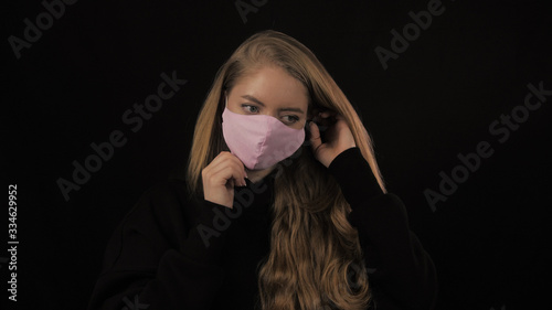 Young woman with golden hair puts on a pink medical mask. Isolated on black background. Health care and medical concept. Close up portrait . 4k. Coronavirus Epidemic  illness  pandemic