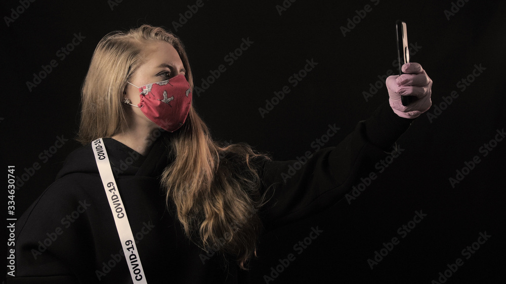 Obraz premium Girl with a white ribbon and the inscription covid 19 in red mask takes selfie on smartphone. Isolated on black background. Health care and medical concept. Coronavirus Epidemic