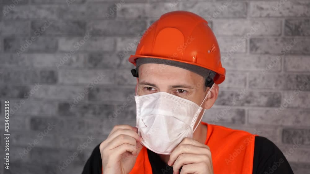 Male construction worker in overalls putting on medical mask on face background of gray brick wall Male construction worker in overalls putting on medical mask on face background of gray brick wall