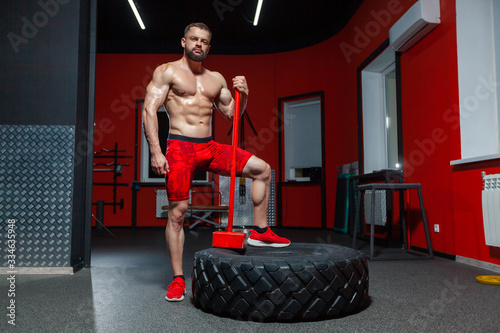 Full length portrait of sporty muscular man posing with sledgehammer and tire in the gym ready to exercise