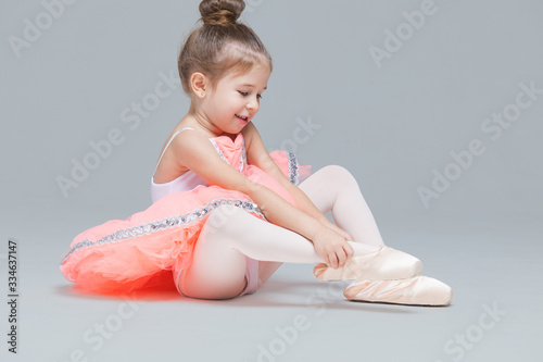 Cute charming ballerina little girl in pink tutu is sitting on the floor trying to put on ballet shoes photo