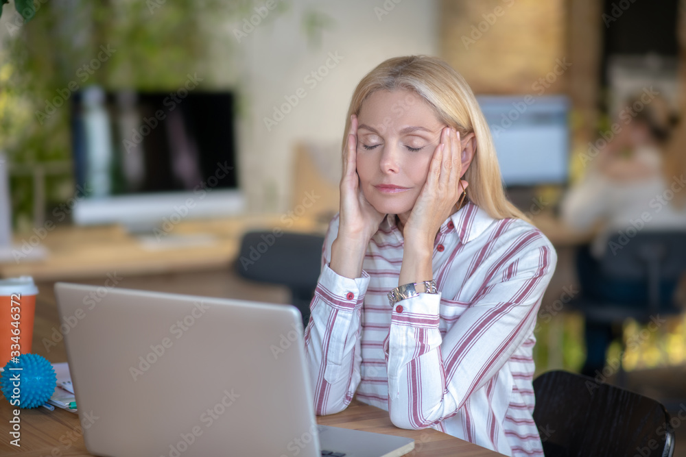 Blonde woman sitting at desk, holding temples Blonde woman sitting at desk, holding temples